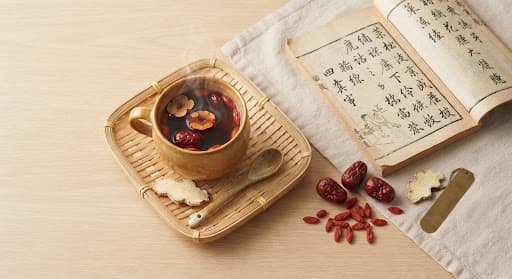 A steaming cup of red date and goji berry tea beside loose red dates and dried ginger